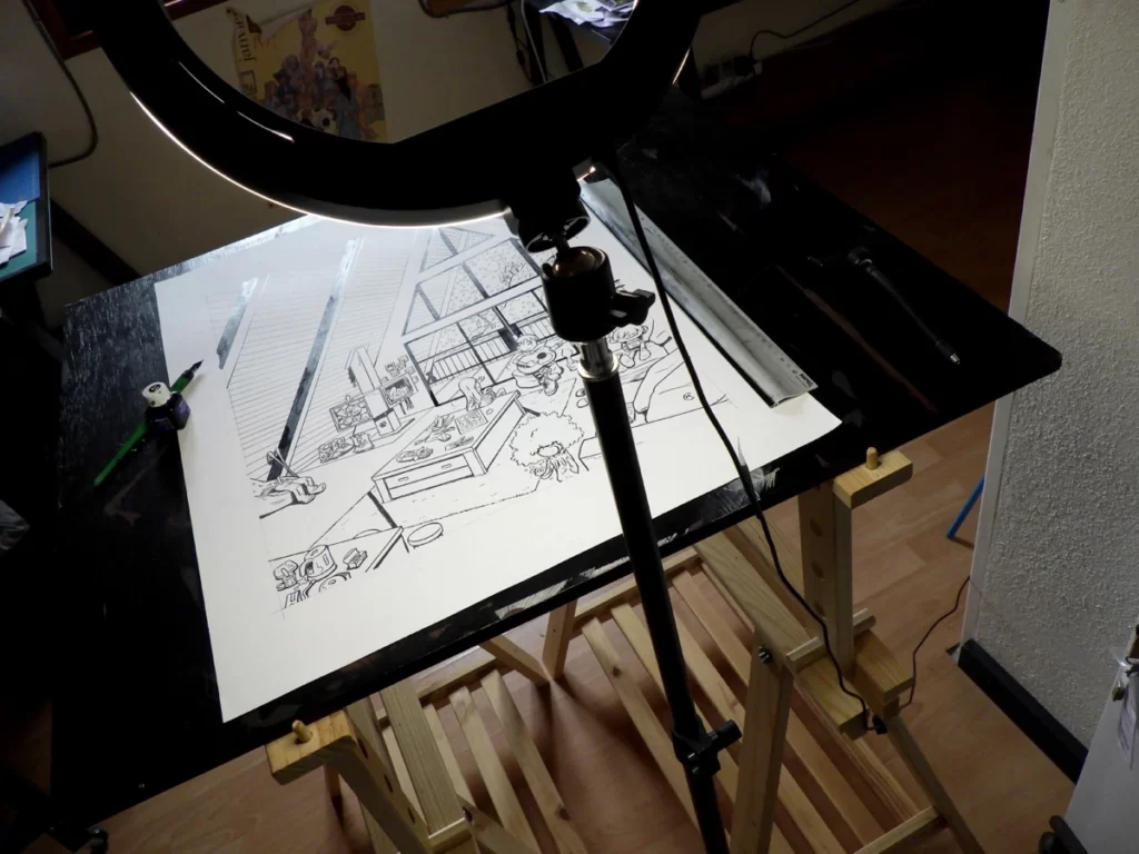Coulisses de la prise devue du dessin à l'encre de chine de pétaroo et ses amis à la neige. On voit une table à dessin et une lampe d'influenceuse ainsi que du matériel à dessin sur la table.