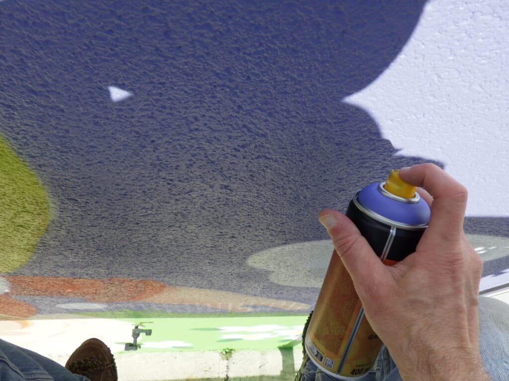 Photo qui montre en gros plan la main de Marcel Champion en train de graffer la fresque Blanche Neige à Nontron. Il utilise une bombe Belton de couleur violette.