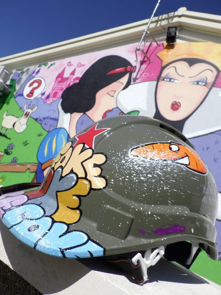 Gros plan du casque de chantier utilisé par Marcel Champion pour réaliser la fresque Blanche Neige à Nontron. Le casque est lui-même customisé et décoré. En arrière plan on voit la fresque terminée.