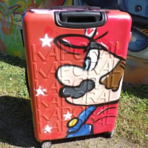 Photo d'une valise rigide à roulette avec la tête de Marion Bros graffée sur un coté. Une belle façon de voyager avec style.
