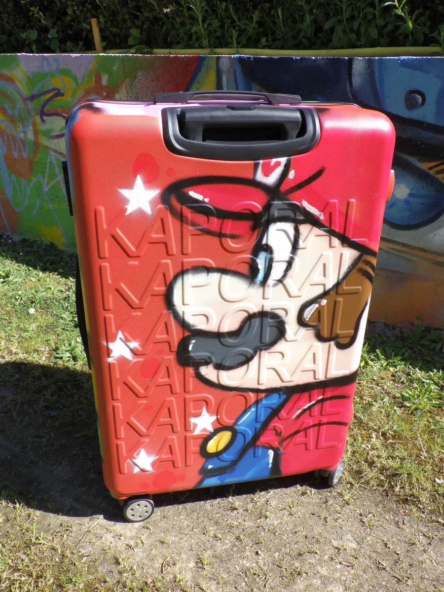 Photo d'une valise rigide à roulette avec la tête de Marion Bros graffée sur un coté. Une belle façon de voyager avec style.