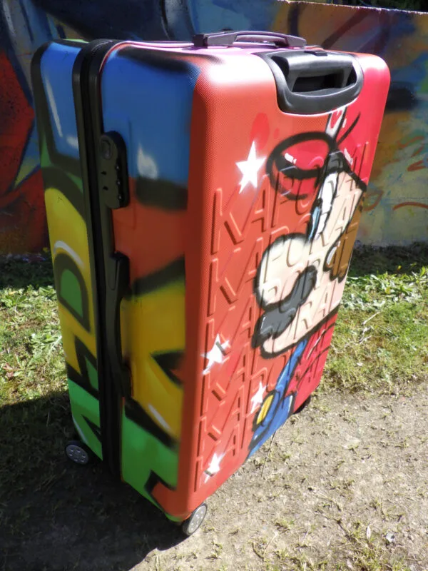 Photo de 3/4 de la valise avec Mario Bros graffé sur une face et on voit sur la tranche de la valise une partie du mot Mario graffé en lettres de couleur. Une belle façon de voyager avec style.