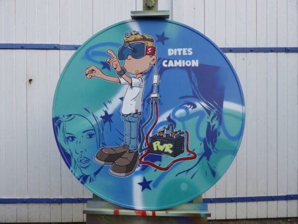 Vue globale d'une peinture réalisée sur un panneau signalétique. Le panneau est de forme ronde. Il s'agissait d'un panneau de limite de hauteur sous pont.On ne voit plus rien des signaux car la totalité a été recouverte. Le fond est peint à la bombe de peinture avec des teintes bleues claires et blanches. Marcel Champion y a ajouté le visages de deux jeunes femmes au pochoir en bleu foncé. Par dessus il a peint un personnage qui utilise un casque de réalité virtuelle fait maison. Il y a même des essuis glace en cas de pluie. Une inscription "dites camion" est présente sur la droite du panneau et fait référence à l'expression "dites camion... Pouet Pouet".