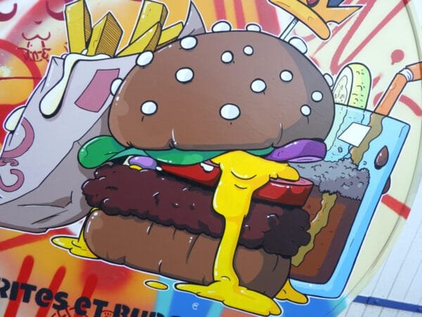 Gros plan sur l'élément principal du premier plan, le burger. On y voit un burger très généreux et bien garni. le fromage a fondu et coule le long du sandwich. Deux tranches de pain entoure un steack généreux et sa garniture. Le burger, les frites et le verre de soda sont réalisés au pinceau contrairement au fond qui est fait à la bombe de peinture.