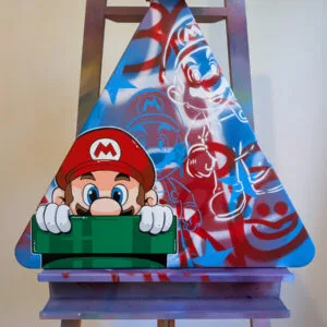 Photo d'une peinture représentant au premier plan Mario sortant d'un tuyau vert. Au second plan on voit un fond graffé avec des pochoirs de mario en blanc et en bleu électrique. Le liseret blanc qui entoure le personnage de Mario permet de le faire ressortir du fond. La peinture est réalise sur un panneau signalétique recyclé, triangulaire.