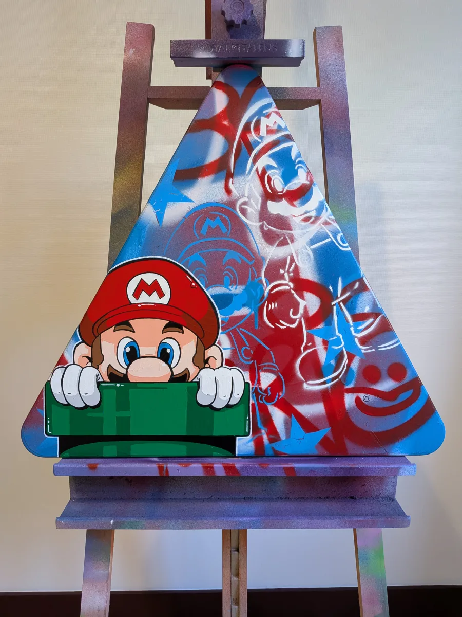 Photo d'une peinture représentant au premier plan Mario sortant d'un tuyau vert. Au second plan on voit un fond graffé avec des pochoirs de mario en blanc et en bleu électrique. Le liseret blanc qui entoure le personnage de Mario permet de le faire ressortir du fond. La peinture est réalise sur un panneau signalétique recyclé, triangulaire.