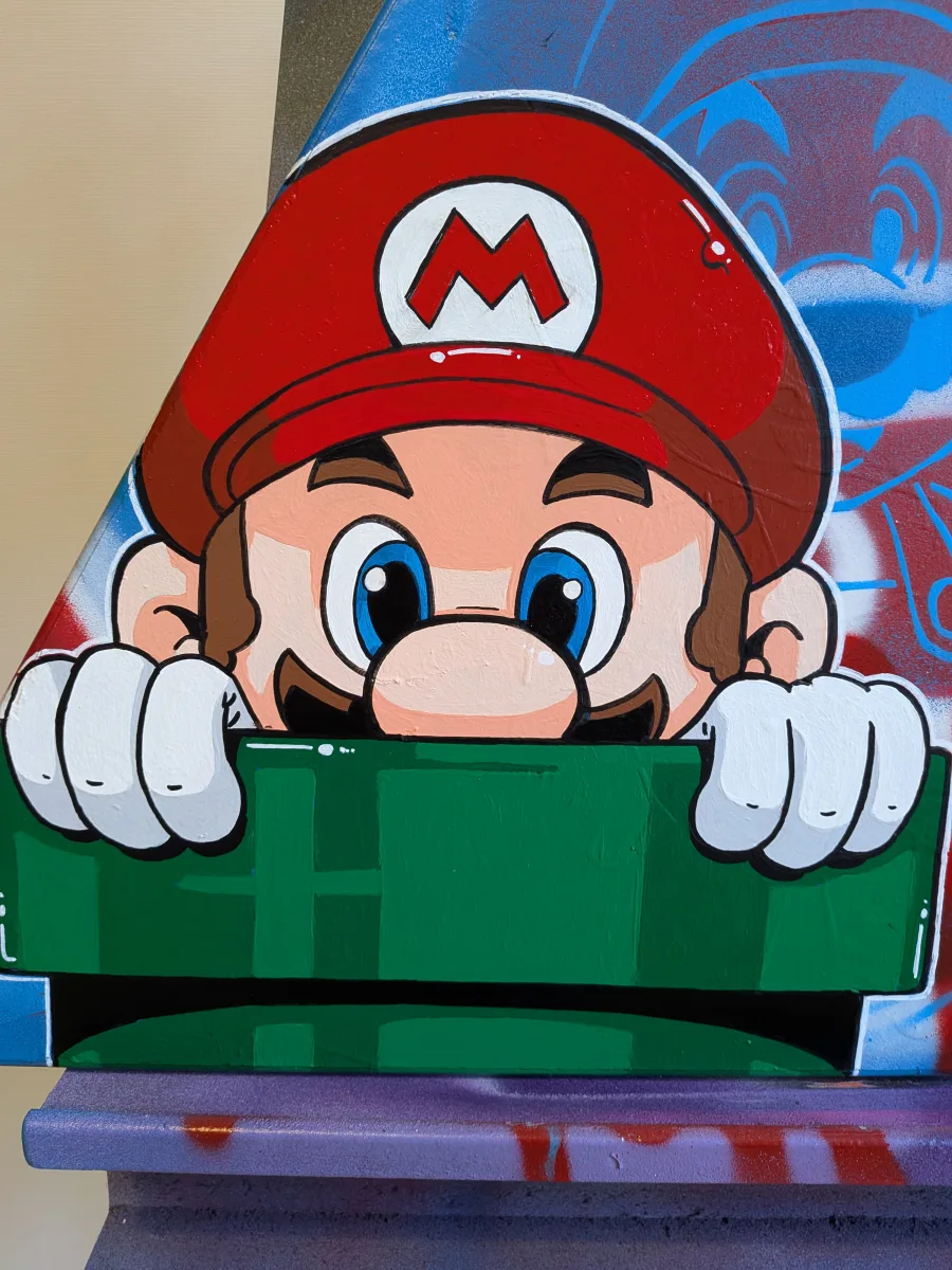 Gros plan sur le Mario en train de sortir d'un tuyau. On voit la partie haute de son visage hors du tuyau et ses deux mains agrippées à ce dernier.