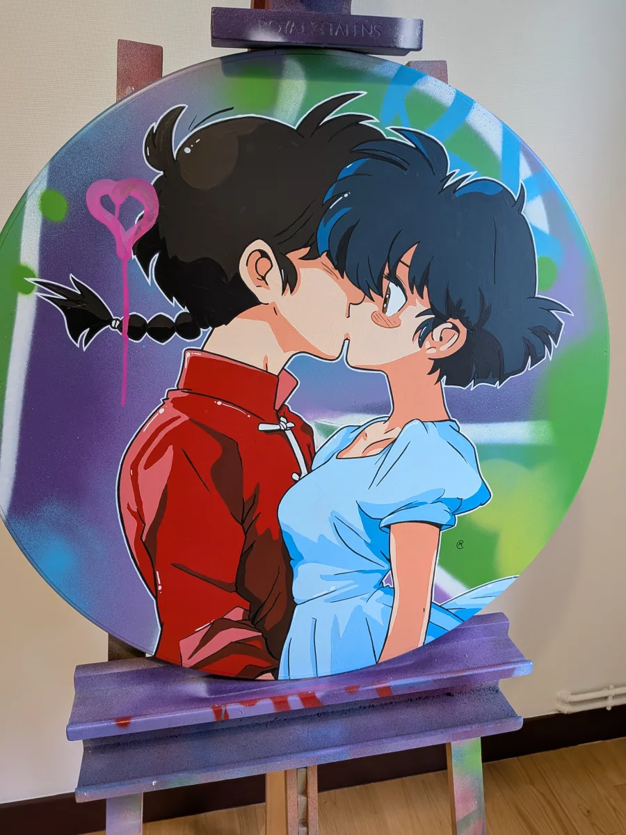 Cette image montre une peinture réalisée sur un authentique panneau de signalisation routière circulaire. L’œuvre représente un couple d’anime emblématique en train de s’embrasser tendrement : une jeune femme aux cheveux courts noirs portant une robe bleu clair, et un jeune homme aux cheveux noirs tressés vêtu d’une tunique rouge traditionnelle chinoise (inspiration claire de Ranma ½ : Ranma Saotome et Akane Tendō). Le fond est traité en street-art coloré avec des dégradés de vert, violet et bleu, et un petit cœur rose tagué ajoute une touche romantique et urbaine. Le panneau conserve ses bords métalliques d’origine et est présenté sur un chevalet éclaboussé de peinture, soulignant le mélange entre signalisation routière détournée et création artistique contemporaine. Une pièce unique qui marie pop culture anime et art de rue sur un support inattendu !