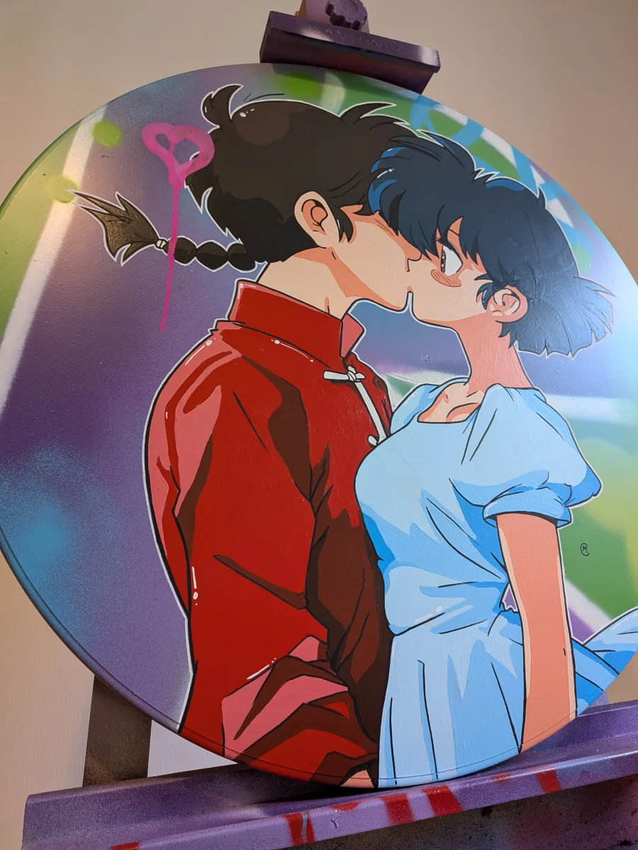Cette image présente une peinture originale réalisée sur un authentique panneau de signalisation routière circulaire. Le couple iconique de Ranma ½ (Ranma en rouge et Akane en robe bleue) s’embrasse passionnément sur un fond street-art aux dégradés violet-vert avec le célèbre cœur rose dégoulinant. L’angle de prise de vue, en plongée légèrement décentrée sur la gauche, crée un effet dynamique : le panneau semble pencher vers nous, comme s’il allait tomber du chevalet. Cette perspective accentue le côté rebelle et vivant de l’œuvre, donnant l’impression que l’amour des deux personnages défie la gravité et sort littéralement du cadre rigide du vieux panneau routier. Une mise en scène audacieuse qui renforce le caractère pop et urbain de la pièce !