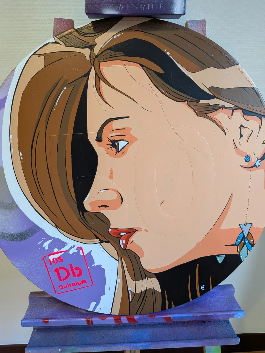 Œuvre street-art ronde sur panneau signalétique authentique : portrait pop-art sensuel d’une pin-up, Dorothy Black, brune aux lèvres rouges pulpeuses et regard de profil, style BD ultra-graphique avec contours noirs parfaits. Clin d’œil génial, la pastille rouge de l’élément chimique DUBNIUM (Db 105) du tableau périodique est intégrée comme un bijou sur son cou. Fond violet métallique avec coulures blanches, finition glossy et format circulaire idéal pour une déco contemporaine. pin-up street art dubnium Db 105, portrait pop art femme tableau périodique, œuvre ronde signalisation routière sexy, art urbain élément chimique 105, peinture contemporaine pin-up Db, déco murale pop art dubnium 2025