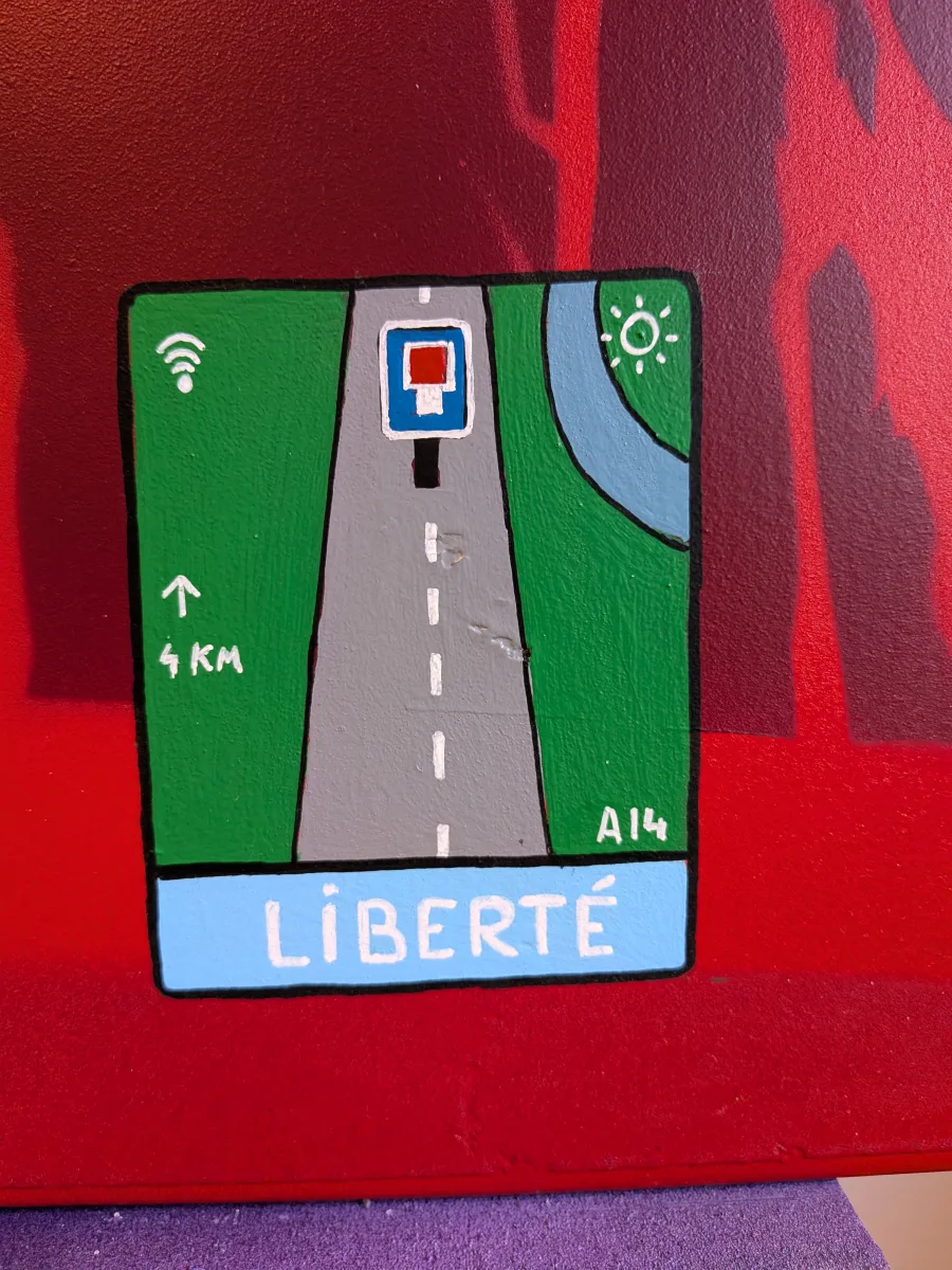 Détail malicieux et génial de l’œuvre street-art sur panneau signalétique : un faux panneau GPS autoroutier peint à la main indique « LIBERTÉ » comme prochaine sortie dans 4 km sur l’A14. Fond vert, icône Wi-Fi, soleil et sens unique renforcent l’illusion parfaite d’un vrai panneau officiel détourné. Sur le fond rouge révolutionnaire de la toile, ce clin d’œil contemporain transforme la Liberté en destination imminente et ironique. Symbole ultime de l’art urbain engagé qui pirate les codes de la signalisation routière. panneau GPS Liberté street art, détournement panneau autoroute A14, sortie Liberté 4 km, art urbain signalisation détournée, Thérébentine détail GPS, street art révolutionnaire français 2025, peinture contemporaine sur panneau routier
