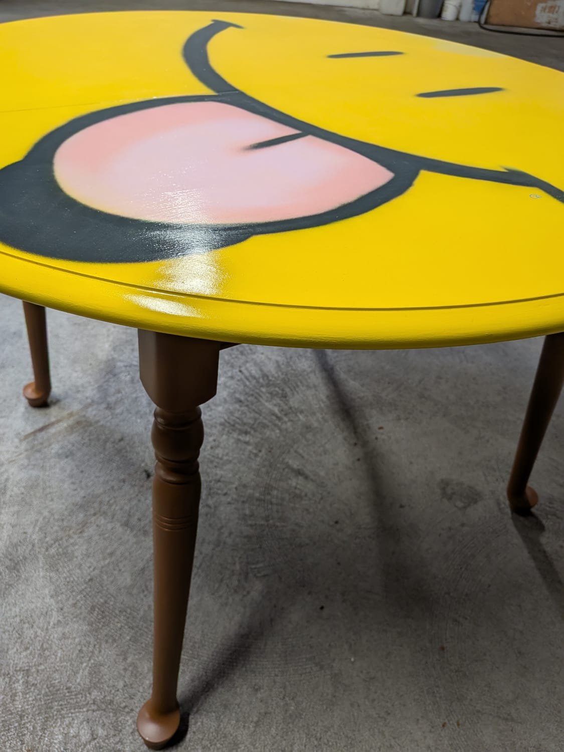 photo d'une table en bois customisée. Le plateau a été peint en jaune vif et représente un emoji qui sourit et tire la langue. La partie basse de la table est peinte en marron qui tire sur le orange.